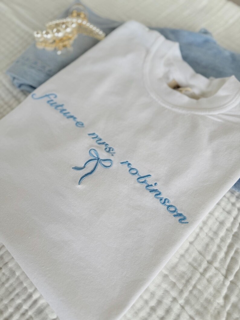 Op de afbeelding: Wit T-shirt met blauwe borduurwerk dat "Future Mrs. Robinson" zegt met een blauwe strik.