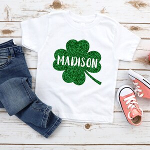 May include: White t-shirt with a green glitter shamrock and the name "MADISON" in white text.