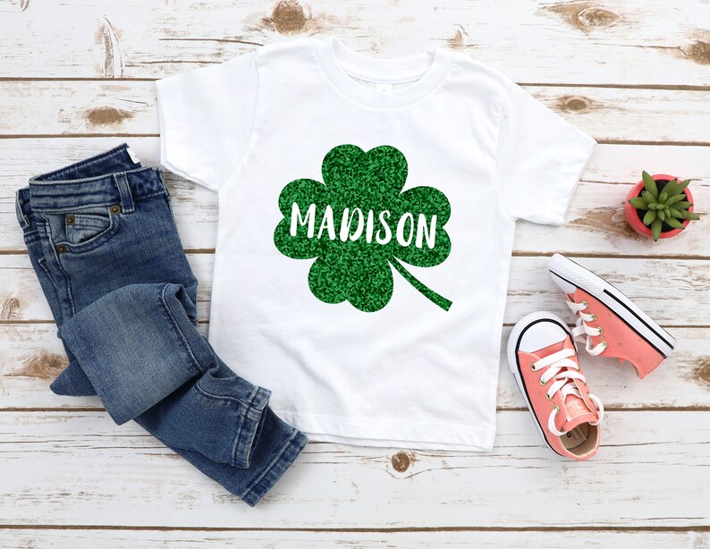 May include: White t-shirt with a green glitter shamrock and the name "MADISON" in white text.