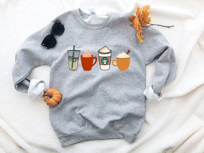 May include: Gray sweatshirt with a graphic of four different coffee drinks, including iced coffee, a latte, a pumpkin spice latte, and a hot chocolate. The sweatshirt is on a white fuzzy surface.