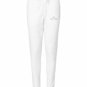 May include: White sweatpants featuring an elastic waistband with a drawstring and the text EST. 08.17.2026 printed in a light brown color on the upper left thigh area. The garment has a tapered leg design with ribbed cuffs at the ankles.