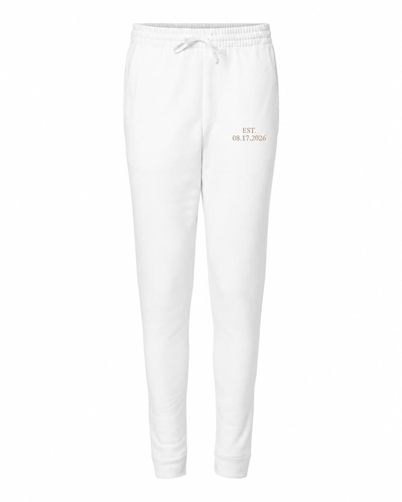 May include: White sweatpants featuring an elastic waistband with a drawstring and the text EST. 08.17.2026 printed in a light brown color on the upper left thigh area. The garment has a tapered leg design with ribbed cuffs at the ankles.