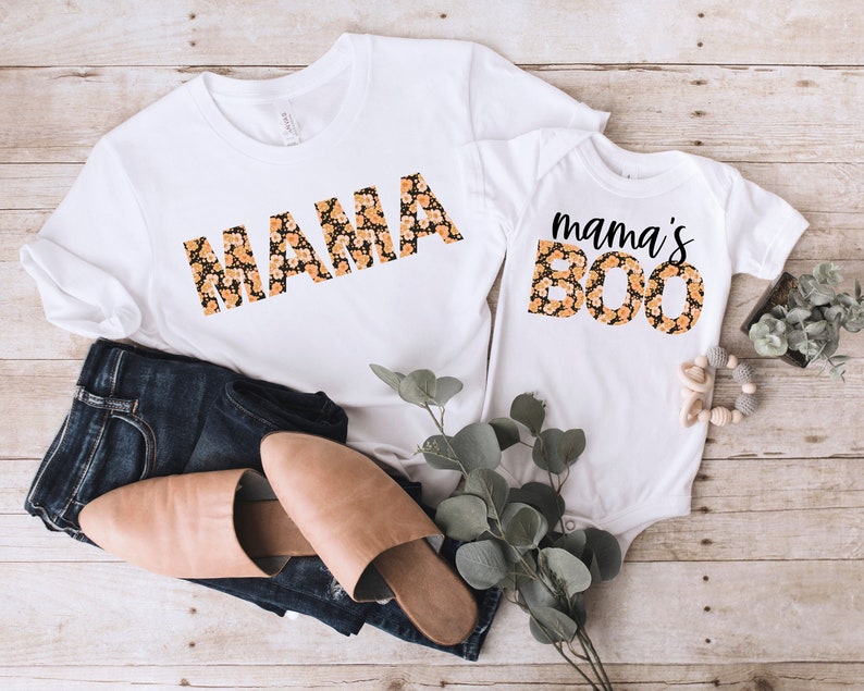 May include: A white t-shirt with the word "MAMA" in black letters with a floral pattern. A white onesie with the text "mama's BOO" in black letters with a floral pattern.