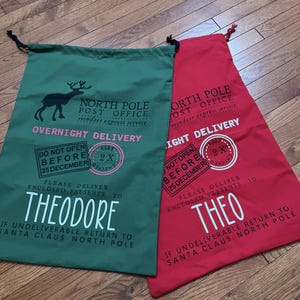 Puede incluir: Dos bolsas de regalo de Navidad, una verde y otra roja, con cordones negros. La bolsa verde presenta un gr&aacute;fico de reno y el nombre "Theodore". La bolsa roja tiene el nombre "Theo". Ambas bolsas tienen texto relacionado con la Oficina de Correos del Polo Norte.