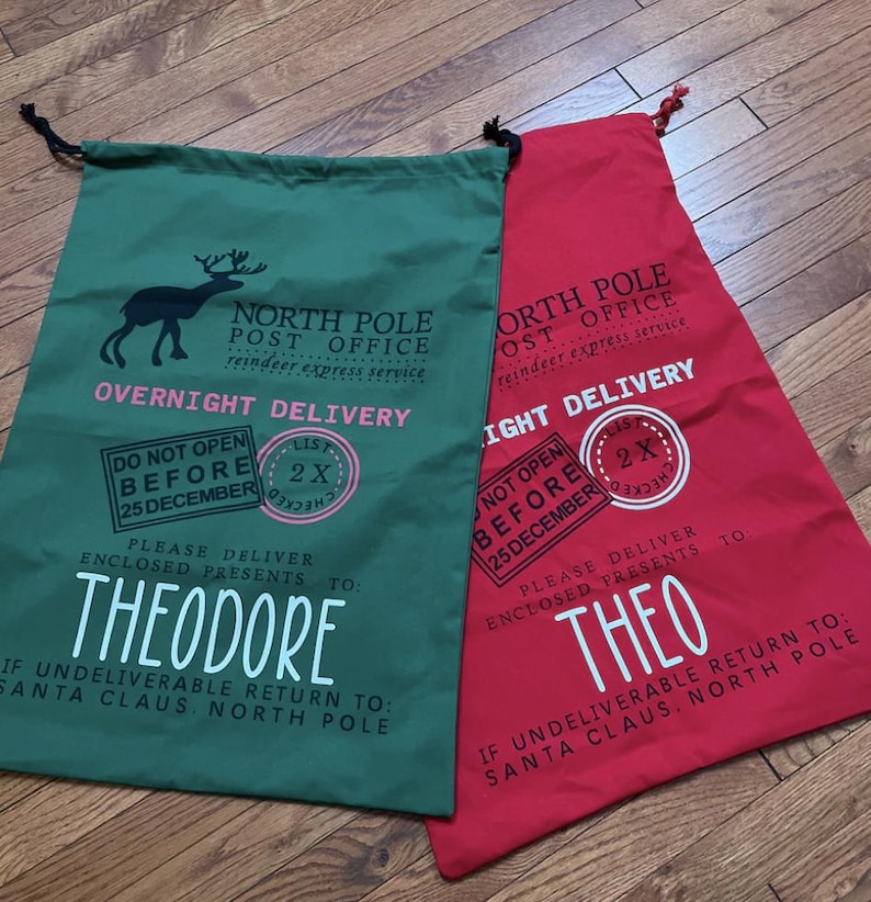 Puede incluir: Dos bolsas de regalo de Navidad, una verde y otra roja, con cordones negros. La bolsa verde presenta un gr&aacute;fico de reno y el nombre "Theodore". La bolsa roja tiene el nombre "Theo". Ambas bolsas tienen texto relacionado con la Oficina de Correos del Polo Norte.