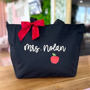May include: Black canvas tote bag with a red bow, the text "Mrs. Nolan" and a red apple graphic.