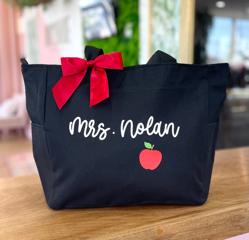 May include: Black canvas tote bag with a red bow, the text "Mrs. Nolan" and a red apple graphic.