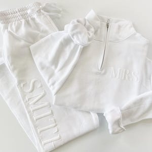 Op de afbeelding: Een witte loungewear set met een joggingbroek met "SMITH" geborduurd op de broekspijp, een sweatshirt met een kwart rits en "MRS" geborduurd op de borst, en een bijpassende scrunchie. De set is gemaakt van een zacht, comfortabel materiaal.