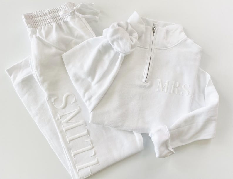 Op de afbeelding: Een witte loungewear set met een joggingbroek met "SMITH" geborduurd op de broekspijp, een sweatshirt met een kwart rits en "MRS" geborduurd op de borst, en een bijpassende scrunchie. De set is gemaakt van een zacht, comfortabel materiaal.