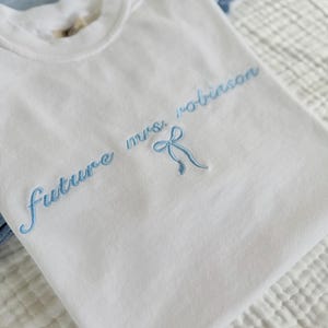 Op de afbeelding: Wit T-shirt met blauwe borduurwerk dat "future mrs. robinson" zegt met een blauwe strik.