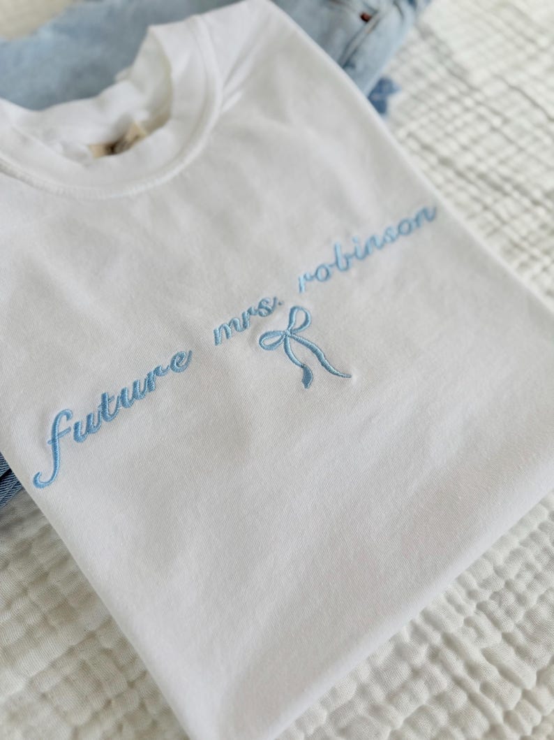 Op de afbeelding: Wit T-shirt met blauwe borduurwerk dat "future mrs. robinson" zegt met een blauwe strik.