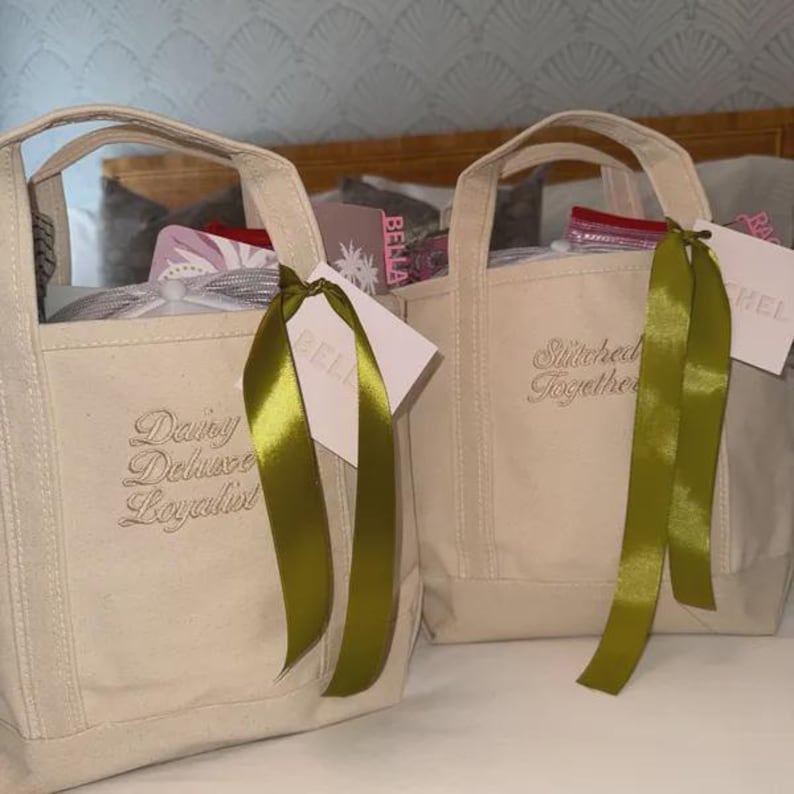 May include: Two off-white canvas tote bags with double handles. The left bag features embroidered text reading "Dairy Deluxe Loyalist" and the right bag features "Stitched Together". Each bag has a green satin ribbon and a white gift tag attached.