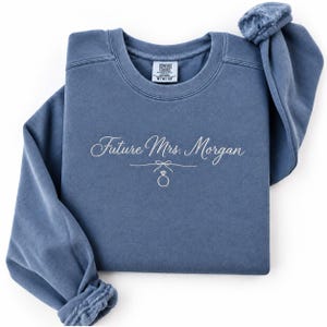 May include: A blue crewneck sweatshirt featuring the embroidered white text Future Mrs. Morgan, accompanied by a small line-art bow and a diamond ring icon. The garment includes a Comfort Colors brand label at the inner neckline showing size M.