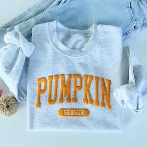 Op de afbeelding: Een lichtgrijs sweatshirt met ronde hals met het woord PUMPKIN in grote, verhoogde, oranje blokletters, met het woord SEASON in een kleiner oranje rechthoekig vlak eronder. Het sweatshirt is gecombineerd met een lichtblauwe spijkerbroek.