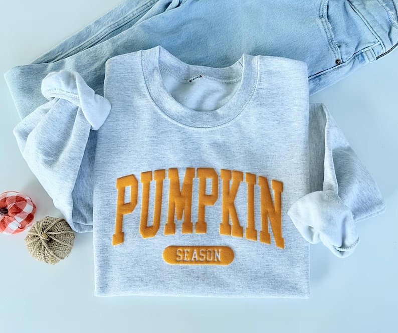 Op de afbeelding: Een lichtgrijs sweatshirt met ronde hals met het woord PUMPKIN in grote, verhoogde, oranje blokletters, met het woord SEASON in een kleiner oranje rechthoekig vlak eronder. Het sweatshirt is gecombineerd met een lichtblauwe spijkerbroek.