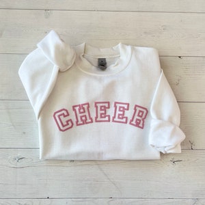 May include: White sweatshirt with a pink "CHEER" graphic in a collegiate font.