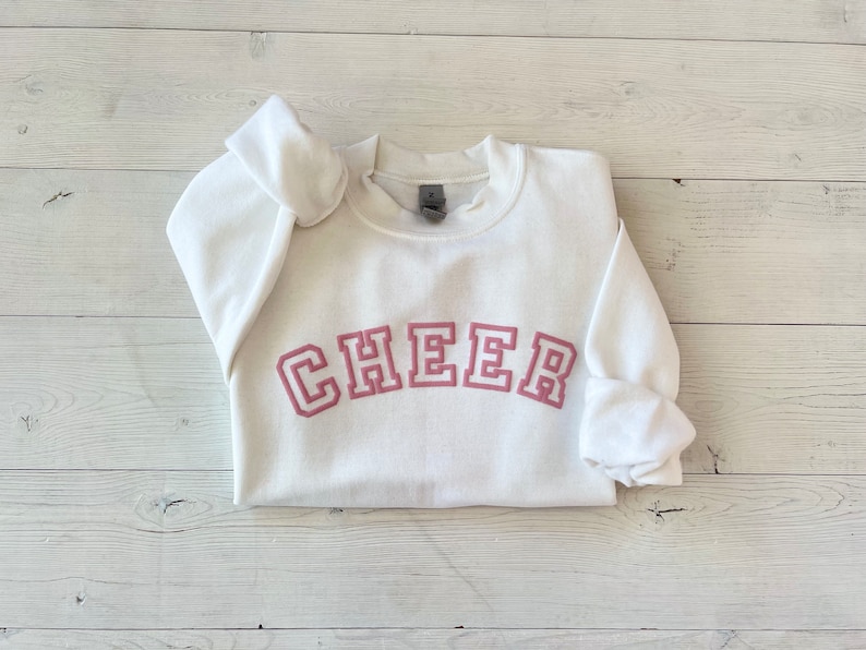 May include: White sweatshirt with a pink "CHEER" graphic in a collegiate font.