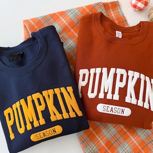 Op de afbeelding: Twee sweaters met ronde hals, &eacute;&eacute;n marineblauw met gele tekst en &eacute;&eacute;n roestoranje met witte tekst. Beide hebben het woord PUMPKIN in grote blokletters boven het woord SEASON in een afgerond rechthoekig kader op de borst.