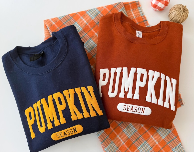 Op de afbeelding: Twee sweaters met ronde hals, &eacute;&eacute;n marineblauw met gele tekst en &eacute;&eacute;n roestoranje met witte tekst. Beide hebben het woord PUMPKIN in grote blokletters boven het woord SEASON in een afgerond rechthoekig kader op de borst.