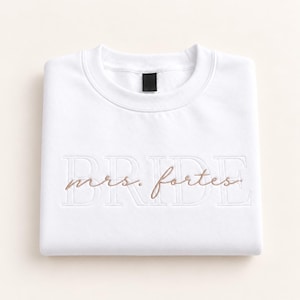 May include: A white crewneck sweatshirt featuring the word BRIDE embroidered in large white block letters across the chest. Overlaid on the block letters is the text mrs. fortes embroidered in a smaller, cursive, light brown thread.