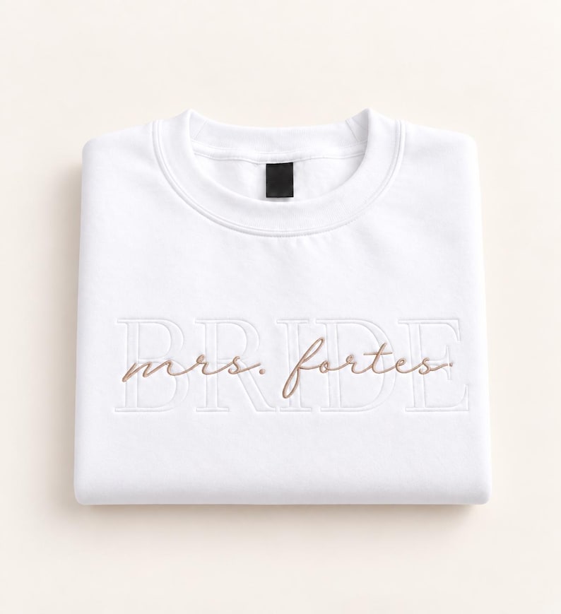 May include: A white crewneck sweatshirt featuring the word BRIDE embroidered in large white block letters across the chest. Overlaid on the block letters is the text mrs. fortes embroidered in a smaller, cursive, light brown thread.