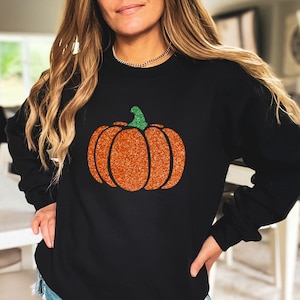 May include: Black sweatshirt with a glittery orange pumpkin design. The pumpkin has a green stem.