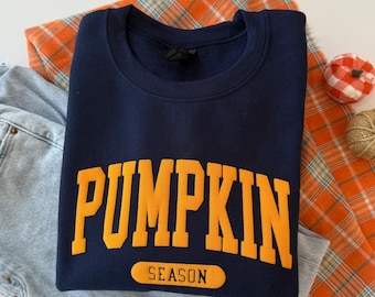 May include: A navy blue crewneck sweatshirt featuring the word PUMPKIN in large, bold, yellow-orange block letters across the chest, with the word SEASON printed in smaller black letters inside a yellow-orange horizontal pill-shaped graphic.