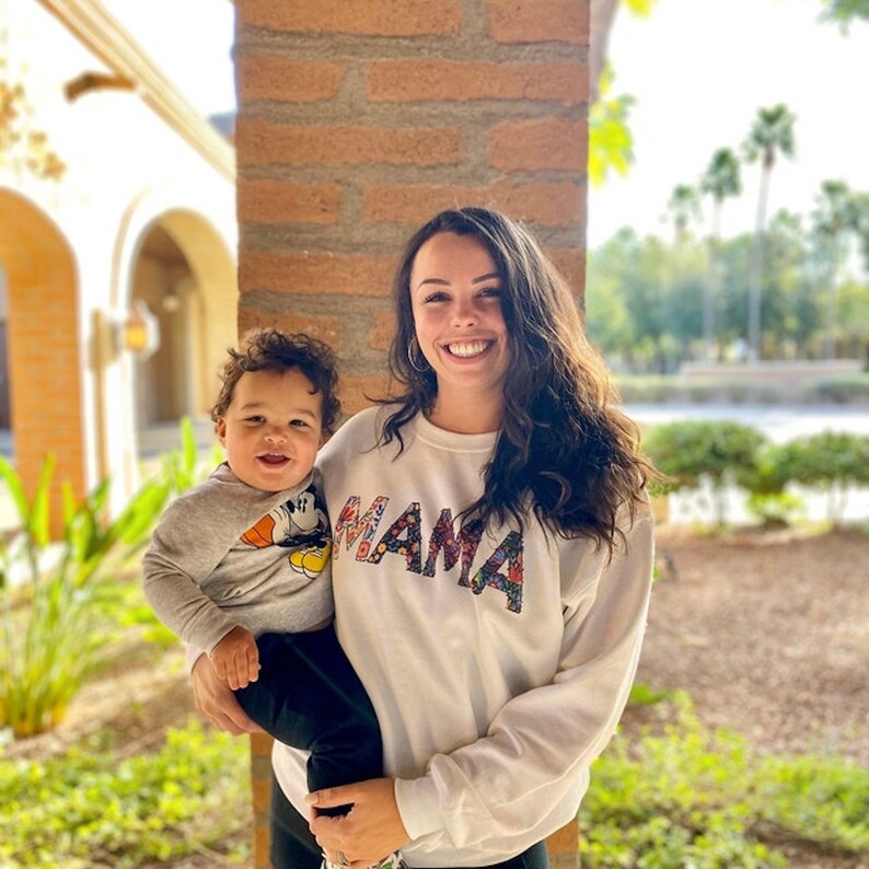May include: A woman wearing a white sweatshirt with the word "MAMA" spelled out in colorful patterned letters is holding a young child. The child is wearing a grey shirt with a Mickey Mouse graphic.