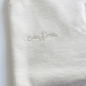 K&ouml;nnte beinhalten: Ein hellbeiger Pullover mit dem Wort "custom" in wei&szlig;er Stickerei.
