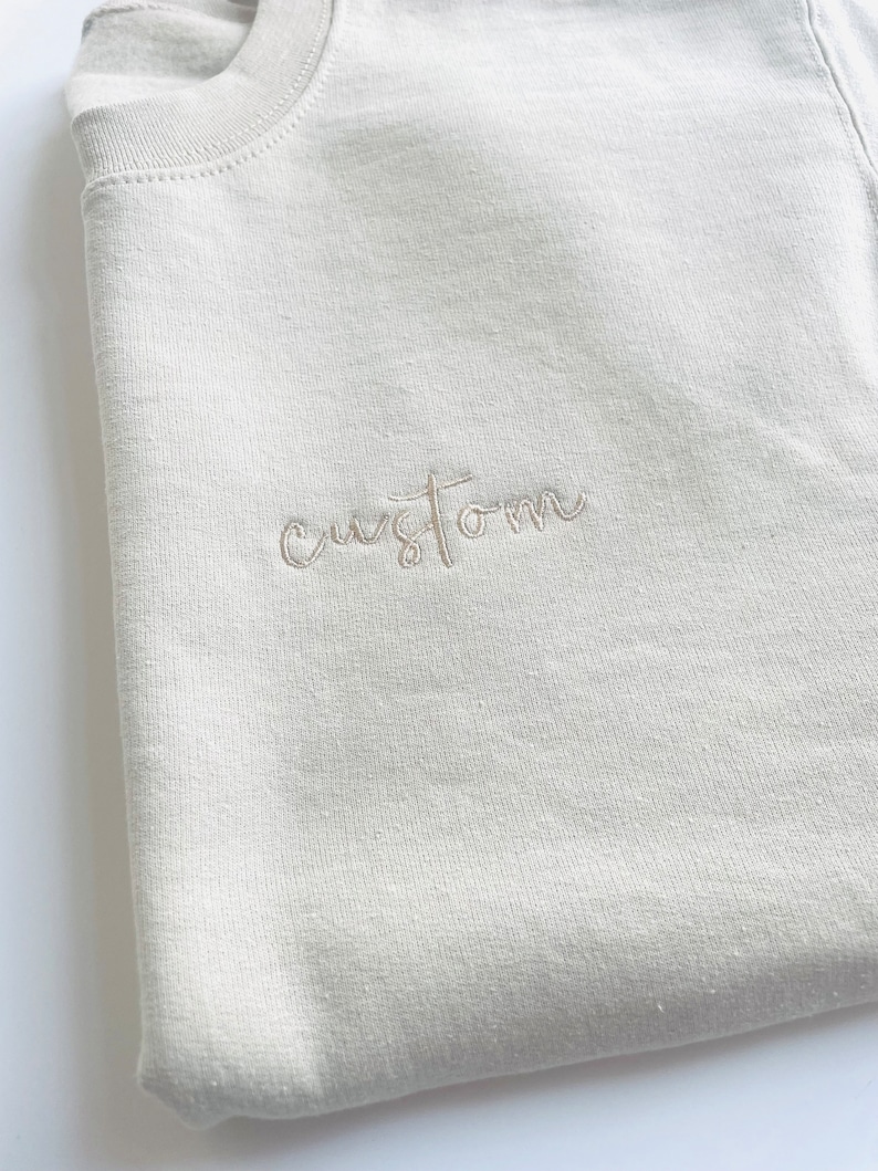 K&ouml;nnte beinhalten: Ein hellbeiger Pullover mit dem Wort "custom" in wei&szlig;er Stickerei.