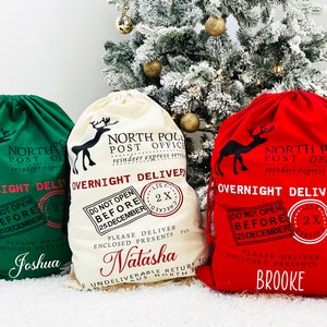 Puede incluir: Bolsas de regalo navide&ntilde;as con cord&oacute;n en verde, crema y rojo. Cada bolsa presenta una silueta de reno, el texto "North Pole Post Office" y "Overnight Delivery". Los nombres est&aacute;n impresos en las bolsas. Las bolsas son de tela.