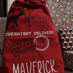 Puede incluir: Un gran saco rojo de Navidad con cierre de cord&oacute;n. Presenta una silueta de reno negra y texto blanco: "North Pole Post Office" y "Overnight Delivery". Un sello dice "Do Not Open Before 25 December". El saco est&aacute; lleno de regalos.