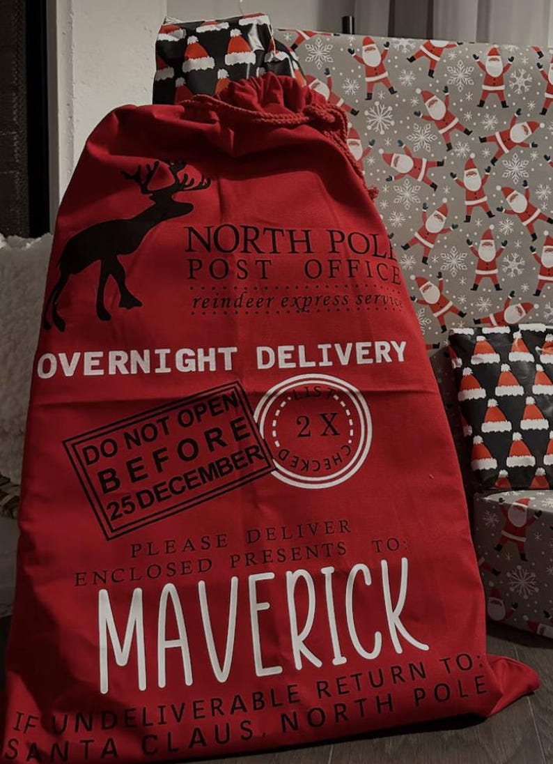 Puede incluir: Un gran saco rojo de Navidad con cierre de cord&oacute;n. Presenta una silueta de reno negra y texto blanco: "North Pole Post Office" y "Overnight Delivery". Un sello dice "Do Not Open Before 25 December". El saco est&aacute; lleno de regalos.