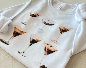 May include: White sweatshirt with a graphic of six different coffee drinks in martini glasses. The coffee drinks are illustrated in a watercolor style.