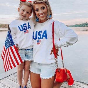 Puede incluir: Una madre y su hija llevan sudaderas blancas con la palabra "USA" impresa en azul con un peque&ntilde;o gr&aacute;fico de bandera estadounidense. Ambas llevan pantalones cortos de mezclilla y sonr&iacute;en a la c&aacute;mara. La madre lleva un bolso rojo y la hija lleva una peque&ntilde;a bandera estadounidense.