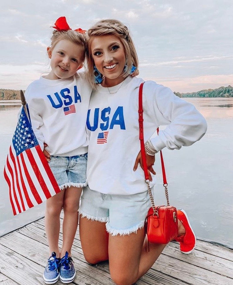Puede incluir: Una madre y su hija llevan sudaderas blancas con la palabra "USA" impresa en azul con un peque&ntilde;o gr&aacute;fico de bandera estadounidense. Ambas llevan pantalones cortos de mezclilla y sonr&iacute;en a la c&aacute;mara. La madre lleva un bolso rojo y la hija lleva una peque&ntilde;a bandera estadounidense.