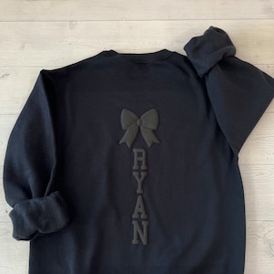 May include: Black sweatshirt with a black bow and the name "RYAN" spelled vertically down the back.