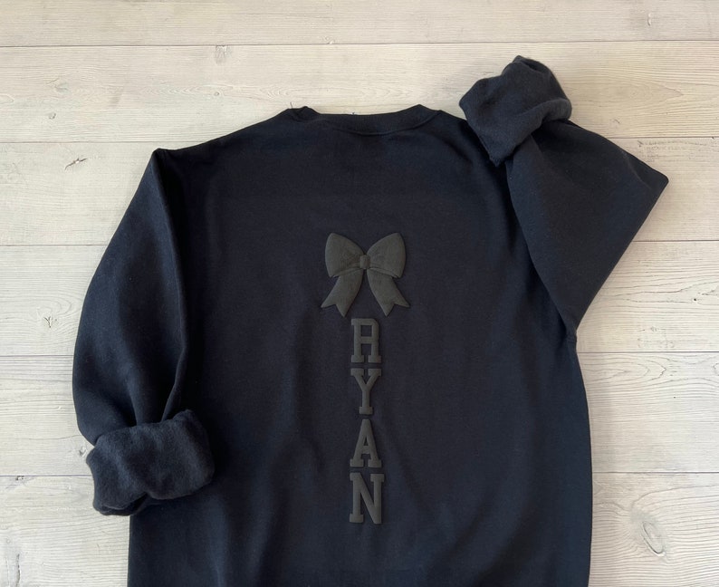 May include: Black sweatshirt with a black bow and the name "RYAN" spelled vertically down the back.