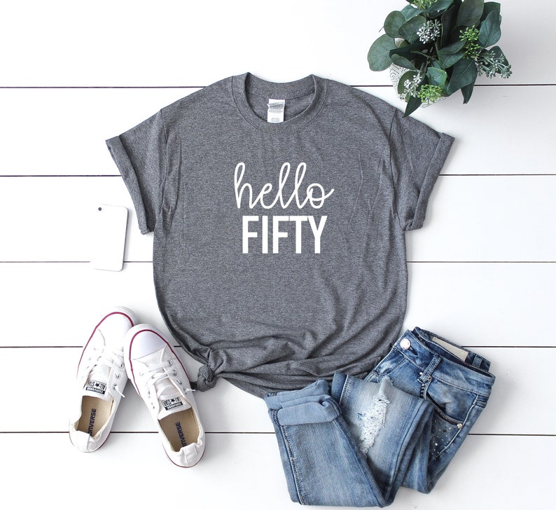 Pode incluir: Camiseta cinza com o texto "hello FIFTY" em letras brancas. A camiseta est&aacute; colocada sobre uma superf&iacute;cie de madeira branca com um par de t&ecirc;nis brancos e uma cal&ccedil;a jeans azul.