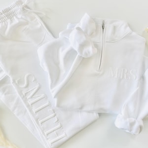 Op de afbeelding: Een witte set met een lange mouwen sweatshirt en joggingbroek. Het sweatshirt heeft een halve rits en de tekst "MRS." geborduurd op de voorkant. De joggingbroek heeft de tekst "SMITH" geborduurd langs de zijkant.