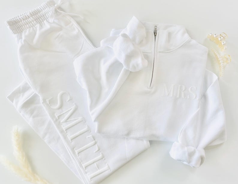 Op de afbeelding: Een witte set met een lange mouwen sweatshirt en joggingbroek. Het sweatshirt heeft een halve rits en de tekst "MRS." geborduurd op de voorkant. De joggingbroek heeft de tekst "SMITH" geborduurd langs de zijkant.