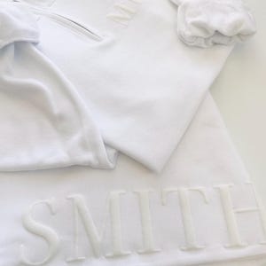Op de afbeelding: Witte trui met een rits en het woord "MRS" geborduurd op de voorkant. De trui is gevouwen over een witte rok met het woord "SMITH" geborduurd aan de onderkant.