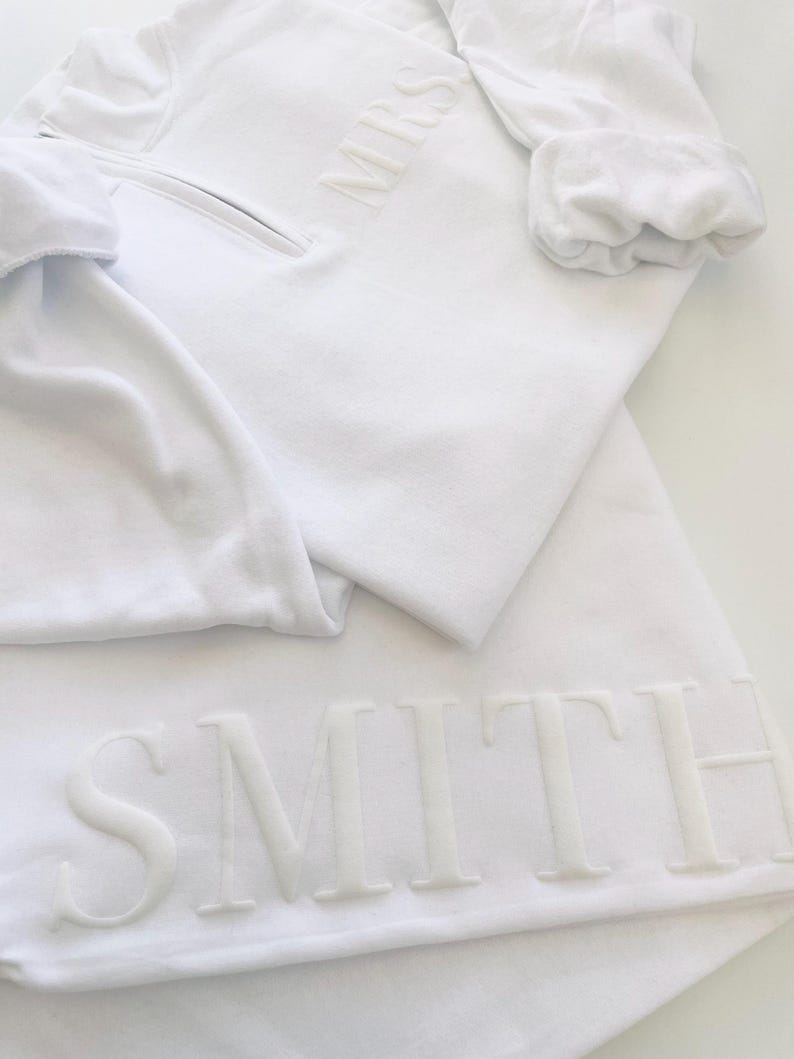 Op de afbeelding: Witte trui met een rits en het woord "MRS" geborduurd op de voorkant. De trui is gevouwen over een witte rok met het woord "SMITH" geborduurd aan de onderkant.