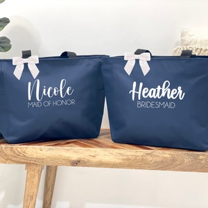 May include: Two navy blue tote bags with white ribbon bows. The bag on the left says "Nicole Maid of Honor" and the bag on the right says "Heather Bridesmaid".
