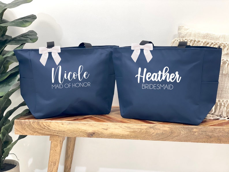 May include: Two navy blue tote bags with white ribbon bows. The bag on the left says "Nicole Maid of Honor" and the bag on the right says "Heather Bridesmaid".