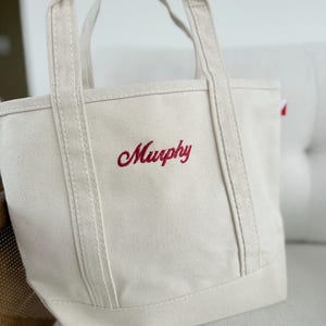 May include: A cream-colored canvas tote bag with red embroidered text that reads "Murphy." The bag has sturdy handles and a rectangular base. The stitching is visible, and the bag appears to be made of a durable material.