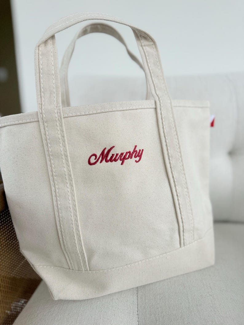 May include: A cream-colored canvas tote bag with red embroidered text that reads "Murphy." The bag has sturdy handles and a rectangular base. The stitching is visible, and the bag appears to be made of a durable material.