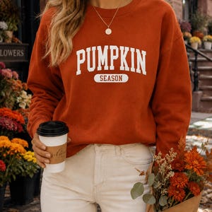 Op de afbeelding: Een oranje-bruine sweater met lange mouwen en een ronde hals, voorzien van de witte tekst PUMPKIN SEASON in een collegiaal lettertype op de borst. Het kledingstuk heeft een geribbelde halslijn en boorden voor een standaard pasvorm.
