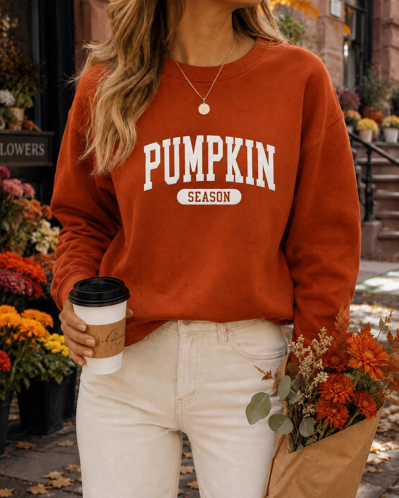 Op de afbeelding: Een oranje-bruine sweater met lange mouwen en een ronde hals, voorzien van de witte tekst PUMPKIN SEASON in een collegiaal lettertype op de borst. Het kledingstuk heeft een geribbelde halslijn en boorden voor een standaard pasvorm.
