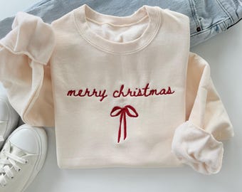 Puede incluir: Sudadera color crema con la inscripción bordada "merry christmas" en rojo, junto con un lazo rojo. La sudadera está doblada, con las mangas visibles. Un par de zapatillas blancas y unos vaqueros azules están en el fondo.